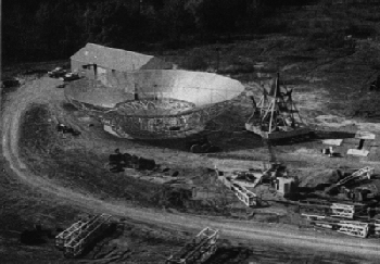 An aerial view of the construction site in October 1969, with the partially finished dish&nbsp;set on the ground next to the base pedestal of four triangular sides. In the foreground are elements of the guy&nbsp;derrick.