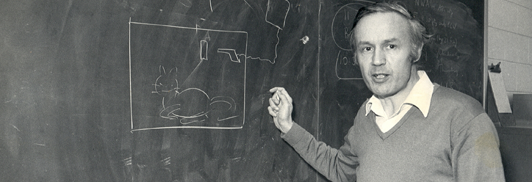托尼·莱格特（Tony Leggett）站在施罗辛格思想实验的手绘插图旁边（注意猫和枪）。